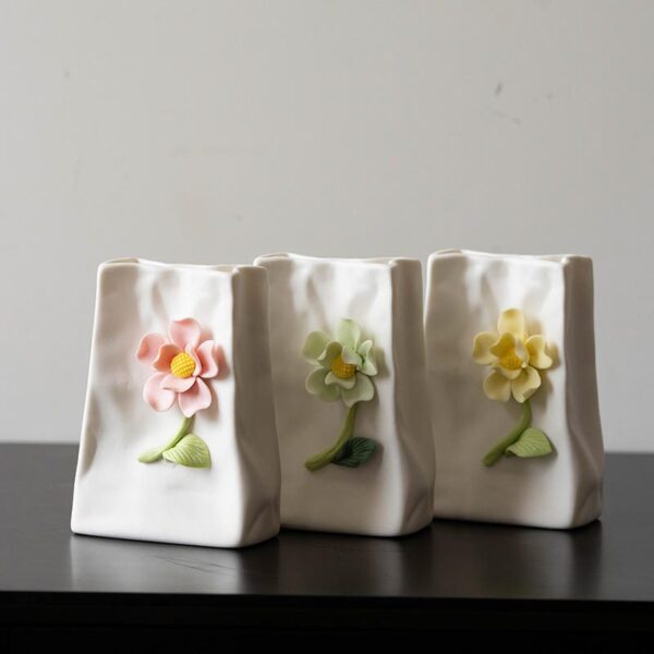 A set of three cute healing-style white ceramic vases, each with a 3D hand-sculpted flower decoration in pink, light green, and yellow, featuring a subtle wrinkled texture. Displayed on a black tabletop against a neutral background, they’re ideal for small fresh flower arrangements or coordinated desktop decor.