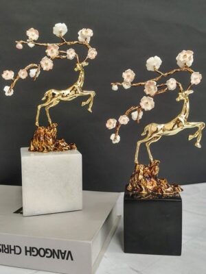 A pair of light luxury decorative ornaments featuring polished brass deer sculptures in leaping poses, paired with enamel plum blossom branches, mounted on white and black marble bases respectively. Displayed against a dark backdrop with art books, the design blends traditional Chinese auspicious symbolism with modern craftsmanship, ideal for modern luxury, new Chinese, or minimalist interiors