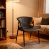 A modern minimalist dining chair upholstered in sleek black faux leather, featuring a curved ergonomic backrest and slim black metal legs. Displayed in a cozy, warmly lit living room with a floor lamp, bookshelf, and sofa, it blends contemporary style with inviting comfort
