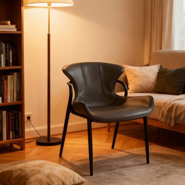 A modern minimalist dining chair upholstered in sleek black faux leather, featuring a curved ergonomic backrest and slim black metal legs. Displayed in a cozy, warmly lit living room with a floor lamp, bookshelf, and sofa, it blends contemporary style with inviting comfort