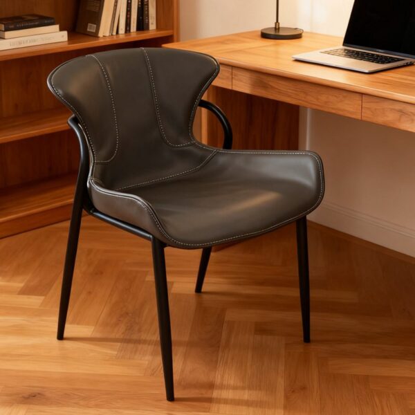 A modern minimalist chair upholstered in black faux leather with contrast white stitching, featuring an ergonomic curved backrest and slim black metal legs. Displayed in a warm home office scene with a wooden desk and bookshelf, it blends style and comfort for dining or workspace use
