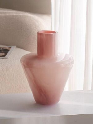 A modern minimalist glass vase with a soft pink gradient finish, featuring a sleek, tapered silhouette and a tall, narrow neck. Displayed on a white tabletop in a sunlit living room with a boucle sofa and sheer curtains, it adds a gentle, elegant focal point to the space
