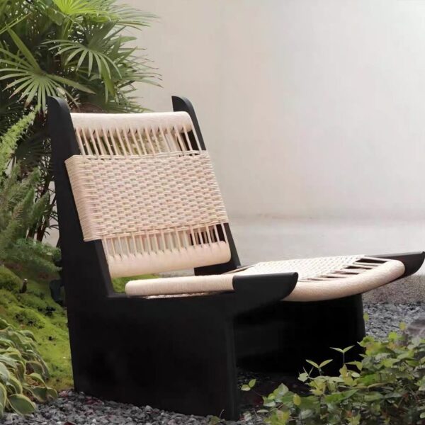 A wabi-sabi style lounge chair with a weather-resistant black solid wood frame and handwoven natural rope upholstery, displayed in an outdoor garden with lush greenery and gravel. It blends rustic texture with minimalist design, ideal for serene outdoor relaxation spaces.