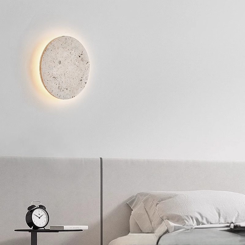 Travertine disc wall sconce mounted above a bed headboard in a minimalist bedroom